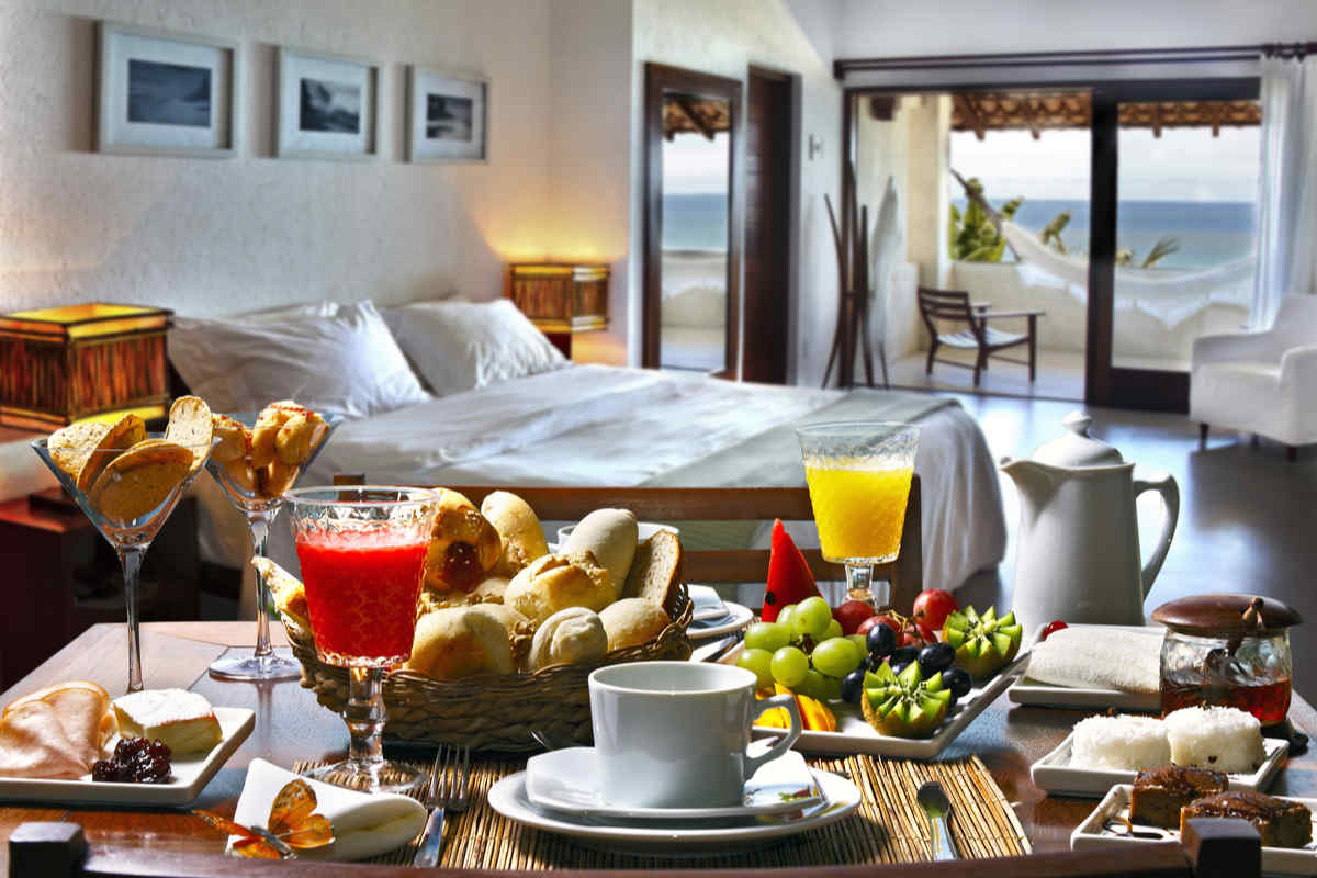 tavolo con vassoio per la prima colazione, con biscotti vari e frutta di vario genere, sullo sfondo letto con lenzuola bianche e finestrone 