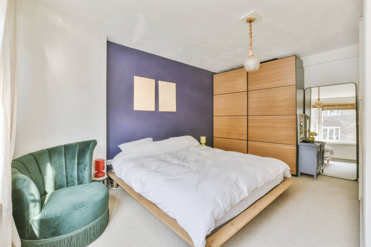 zona notte con letto di fronte a parete di colore viola, poltrona verde, armadio con ante scorrevoli di colore legno e specchio