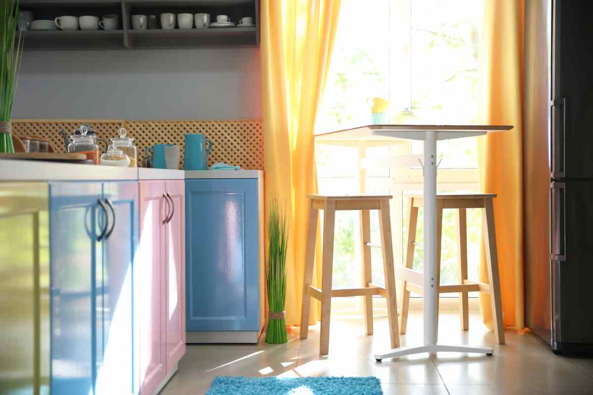 cucina con mobili dipinti con colori diversi e tenda gialla sulle finestre