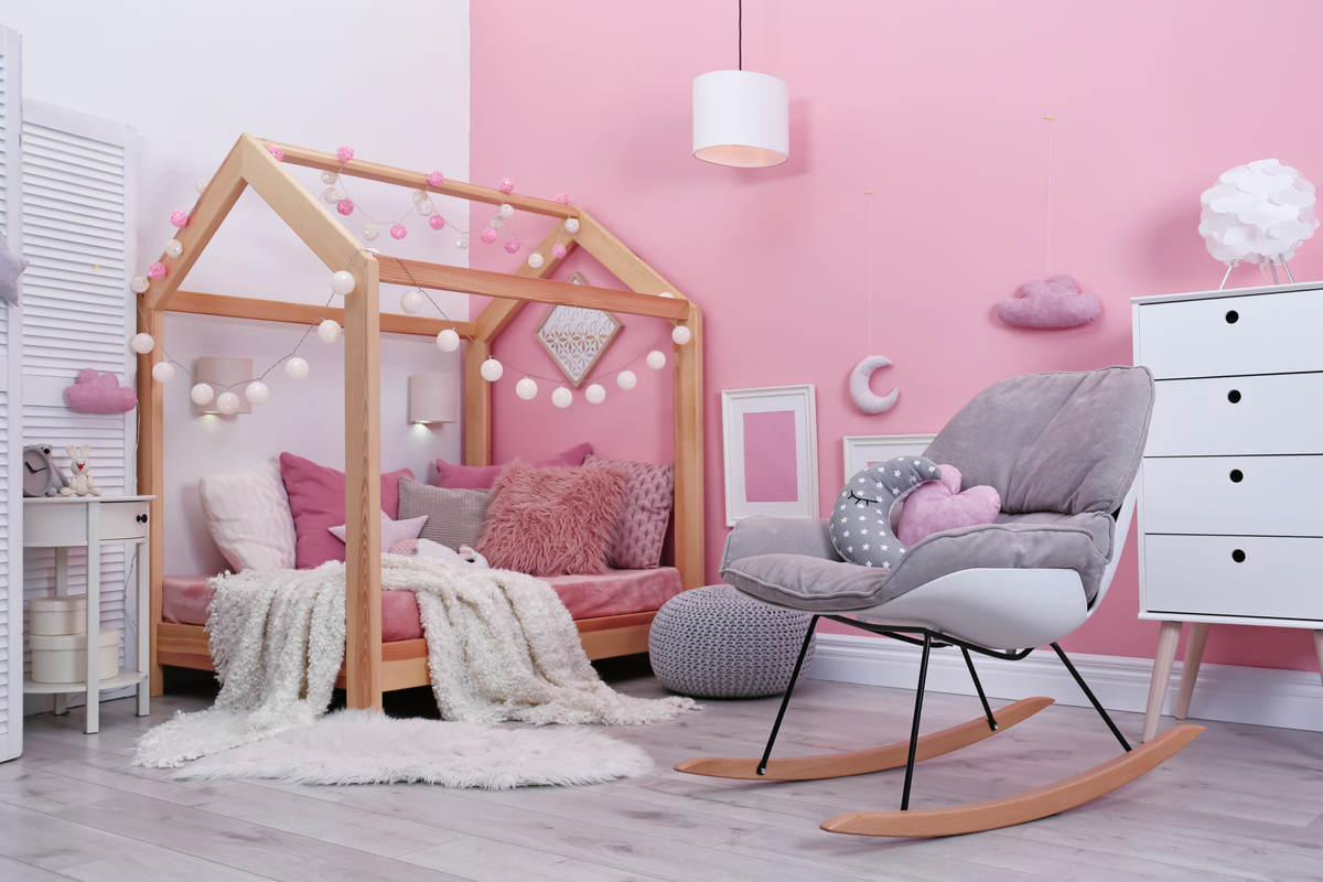 cameretta con parete rosa, letto in legno con lucine appese, sedia a dondolo grigia, cassettiera bianca e pavimento in parquet