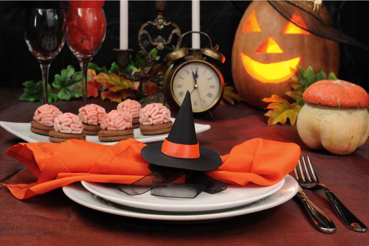 tovagliolo di colore arancione decorato con un cappello da strega su tavola decorata per halloween, sullo sfondo una zucca, una sveglia e dei biscotti decorati con una mousse che riprende le sembianze del cervello