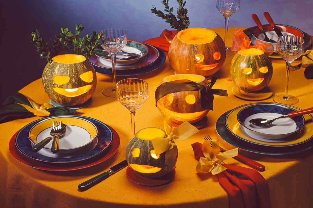 tavola apparecchiata a tema halloween con tovaglia di colore arancione, zucche intagliate, piatte, posate e bicchieri 
