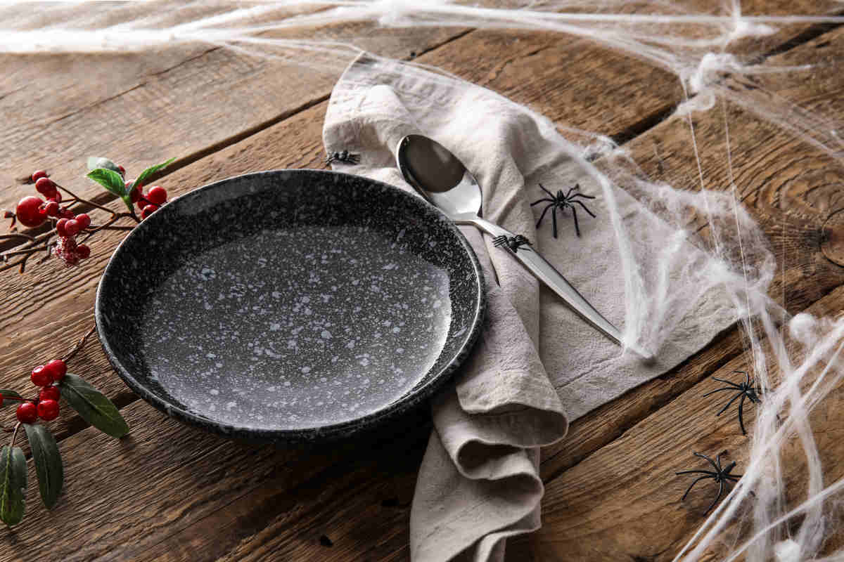 tavolo di legno con ragnatela finta, con piatto nero e cucchiaio poggiato su tovagliolo grigio con ragni finti e ramo di bacche rosse