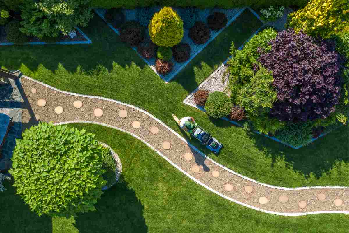 villa con giardino e aiuole fiorite ben curate, cespugli e giardiniere che sta falciando il prato lungo un vialetto visto dall'alto