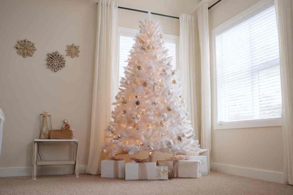 un grande albero di natale bianco con decorazioni in legno e luci led in un angolo del soggiorno davanti alle finestre
