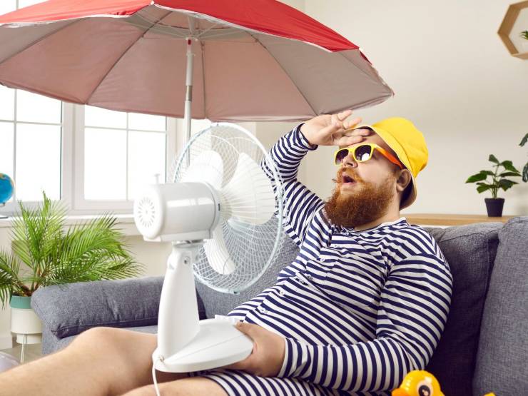 Uomo con ventilatore puntato verso di lui ed ombrellone in casa