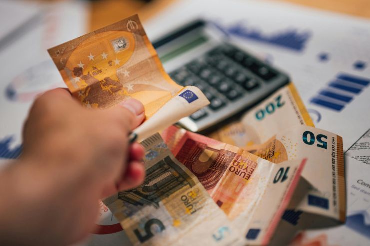 una foto della mano di un uomo che sta pagando una spesa e sullo sfondo si vedono fogli con dei grafici ed una calcolatrice