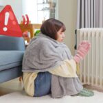 Una ragazza con una coperta e guanti che ha vicino un termosifone ed in alto a sinistra un segnale di pericolo