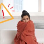 Una ragazza a casa sul divano che sente freddo con una coperta attorno a se ed un segnale di pericolo in alto a sinistra