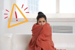 Una ragazza a casa sul divano che sente freddo con una coperta attorno a se ed un segnale di pericolo in alto a sinistra