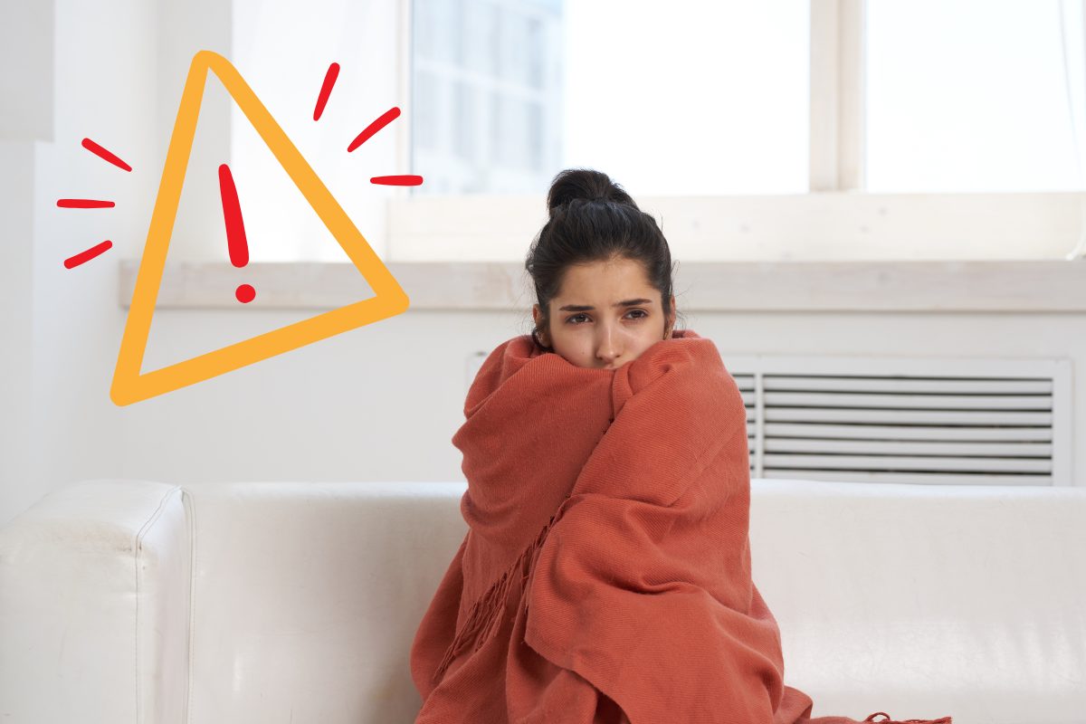 Una ragazza a casa sul divano che sente freddo con una coperta attorno a se ed un segnale di pericolo in alto a sinistra
