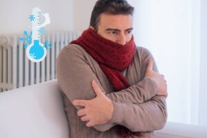Un uomo in casa che sente freddo ed un termometro che raffigura una temperatura ghiacciata, in alto a sinistra