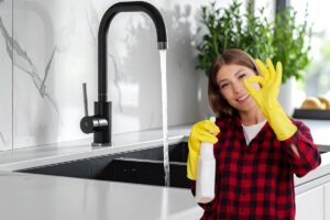 Un rubinetto della cucina nero ed una signora con guanti e prodotti per pulire che fa cenno di "Ok"