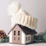 Dei rami e delle pigne sul tavolo vicino ad una miniatura di una casa con sopra un cappello di lana