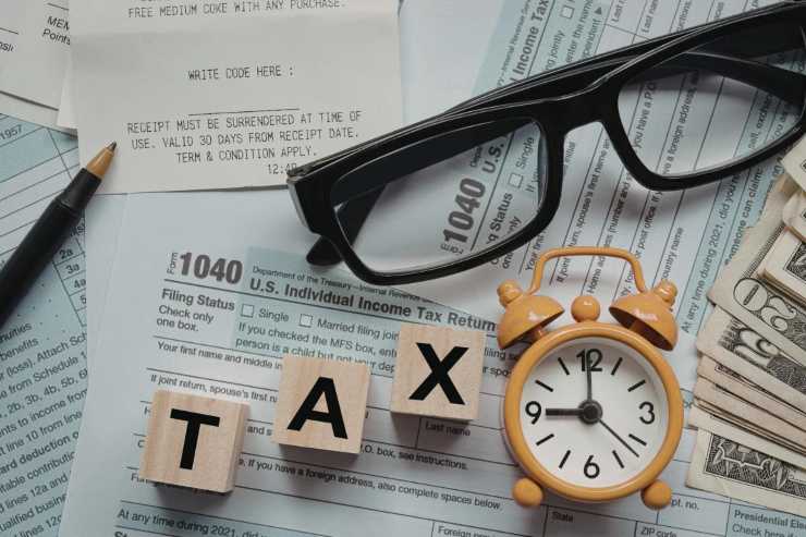 La foto raffigura una composizione che simboleggia la stagione delle imposte. In primo piano, si vedono degli occhiali neri, una sveglia color zucca che segna le 12:00, e quattro blocchi di legno che compongono la parola TAX (tassa). Lo sfondo è costituito da moduli fiscali statunitensi, in particolare il Modulo 1040 (U.S. Individual Income Tax Return), scontrini e banconote, suggerendo la necessità di preparare e pagare le tasse entro la scadenza.