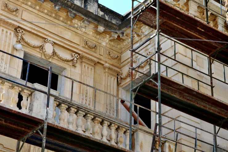 La foto cattura il dettaglio di un edificio storico o antico con una facciata riccamente decorata, caratterizzata da balaustre in pietra lavorata e cornici ornate. La struttura è parzialmente coperta da una robusta impalcatura metallica, indicando lavori di restauro o ristrutturazione in corso.