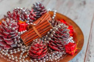 Un dettaglio ravvicinato di una decorazione natalizia, dove pigne dipinte di rosso e naturale, bastoncini di cannella legati insieme, perline argentate e piccole scatoline regalo rosse sono disposti su un vassoio di rame. L'immagine cattura i colori e le texture che richiamano il periodo festivo.