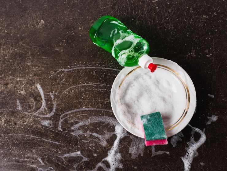 superficie scura su cui sono poggiati un piatto insaponato, del sapone per piatti e una spugnetta