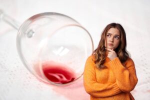 in primo piano una donna che pensa, sullo sfondo una tovaglia chiara macchiata con del vino rosso caduto dal calice
