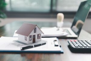 Una miniatura di una casa sopra dei documenti con un computer ed una calcolatrice sulla scrivania