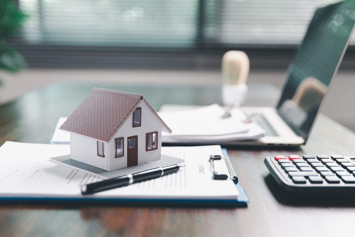 Una miniatura di una casa sopra dei documenti con un computer ed una calcolatrice sulla scrivania