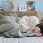 Una persona sdraiata che sente freddo in casa e si copre ed una cornice di fiocchi di neve che rende l'atmosfera ghiacciata