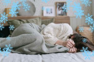 Una persona sdraiata che sente freddo in casa e si copre ed una cornice di fiocchi di neve che rende l'atmosfera ghiacciata