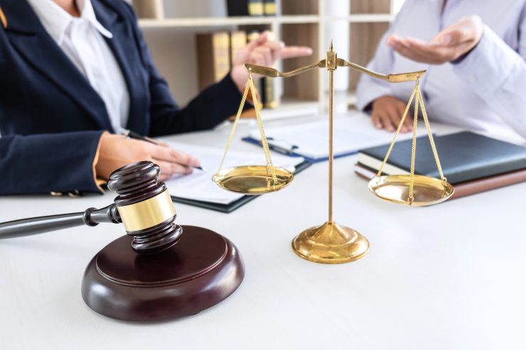 Un tavolo con sopra documenti e una bilancia di giustizi e un martello da giudice, con davanti due persone che discutono