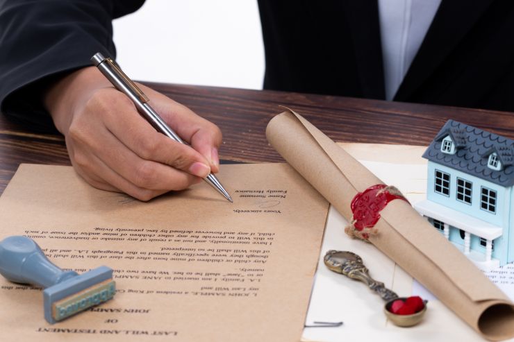 Un erede che firma dei documenti e sopra al tavolo si vede una miniatura di una casa, una pergamena, un cucchiaio ed un timbro.