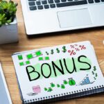 Una scrivania con sopra un computer un mouse e un quaderno con sopra disegnato una scritta "Bonus"