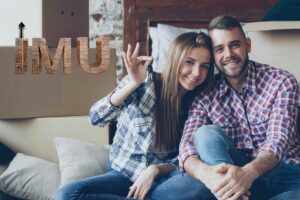 Una coppia felice che ha attorno scatole per il trasloco ed una scritta sulla sinistra fatta in mattoni "IMU"